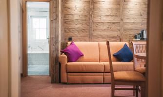 A cozy living room with an orange sofa and colorful cushions. In the background, a bathroom is visible, accessible through a glass door.