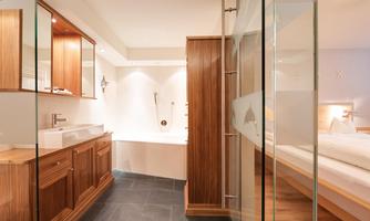A modern bathroom with wooden paneling and an elegant bathtub. There is a large shower and an open living area with a window.