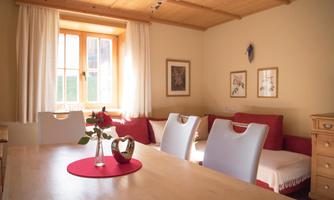 A cozy living space with a bright wooden ceiling and large windows. The table is set and there are flowers in a vase.