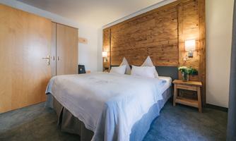 A modern bedroom with a double bed and wooden paneling. The room is cozy and well-lit, with a wardrobe and a nightstand.