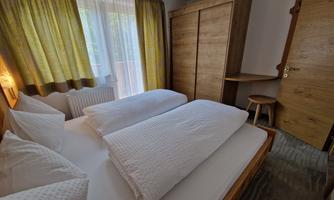 A cozy bedroom with two beds, white bedspreads, and wooden furniture. The room is bright and friendly, with large windows and curtains.