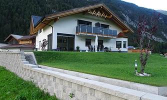 A modern house with wooden details, surrounded by green lawn and mountains in the background. The large terrace offers space to relax outdoors.