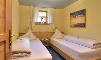 A cozy bedroom with two single beds and warm yellow walls. The room is bright and inviting, with a window view and a painted picture on the wall.