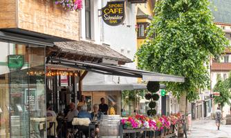 A cozy café with colorful flower boxes and a covered terrace. It's lightly raining, and people are sitting outside relaxed.