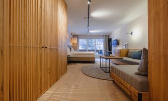 A modern living room with wooden paneling and soft lighting. In the background, you can see a cozy sleeping area with a window and a TV.