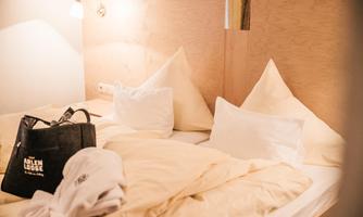 A cozy bedroom with two beds and white pillows. On top lies a bag and a bathrobe.