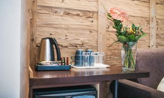 A small table with a kettle, glasses, and a flower vase stands against a wooden wall. Next to the table is a comfortable armchair.