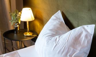 A cozy bed with white pillows and a stylish lamp next to it. In the background, there is a green wall and a small floral decoration.
