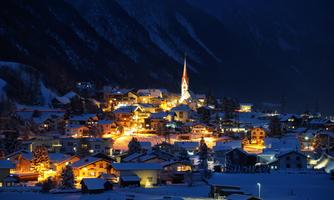 A picturesque village at night, with snow-covered houses and warm lights. In the background, a church tower rises into the clear, dark sky.