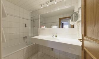 A modern bathroom with white tiles and a bathtub. The area is well-lit and features a large mirror.