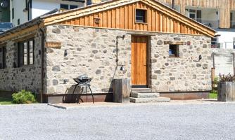 A rustic stone house with a wooden roof and a wooden entrance. Outside there is a barbecue and there is a small garden area.