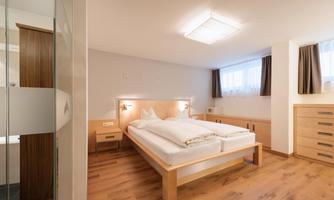 A modern bedroom with a large double bed and light wooden decor. The walls are painted in neutral tones, and there are large windows with curtains.