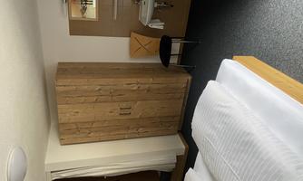 A simple hotel room with two beds, a wooden wardrobe, and a small sink. The room has a neutral color tone and carpeted flooring.