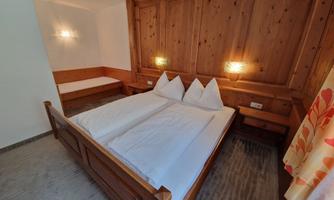 A cozy bedroom with a large double bed and wood paneling. Next to it is a single bed and soft lighting.