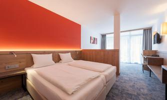 A modern hotel room with two single beds and a warm red wall. Large windows provide plenty of daylight and offer access to a balcony.