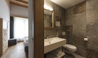 A modern bathroom with gray tiles and a clear mirror. In the background, a bright bedroom with a window is visible.