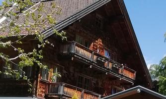A traditional chalet with wooden cladding and balconies. The building is located in a green environment with a wooden fence in the foreground.