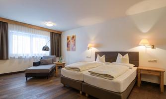 A cozy bedroom with two beds and a modern style. The room features a window, a seating area, and warm lighting.
