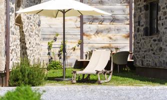 A cozy outdoor area with a lounge chair and a sun umbrella. In the background, stone houses and a green meadow can be seen.