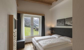 A cozy room with two single beds and a window that looks out onto a green landscape and mountains. The decor is modern and friendly.