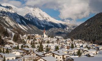 A picturesque winter landscape with snow-covered houses and majestic mountains in the background. The sky is partly cloudy, creating a serene atmosphere.
