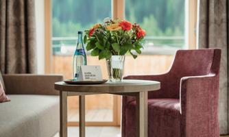 A cozy room with a table on which a flower vase and a water bottle are standing. In the background, there are windows overlooking a green landscape.