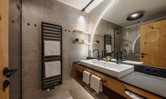 A modern bathroom with a large sink and two faucets. The walls are in neutral colors, and there are towels as well as wooden shelves.