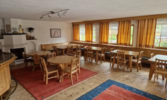 A cozy room with wooden tables and chairs. Large windows with orange curtains let in a lot of light.
