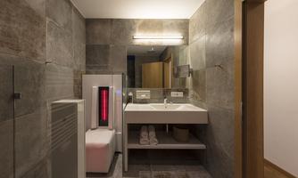 A modern bathroom with gray tiles and bright lighting. There is a double sink, towels, and an infrared sauna.