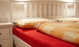 A bed with a white headboard and red bed sheets is placed in a cozy room. The comforter has a warm, yellow pattern and there are nightstands next to the bed.