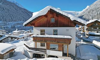 A picturesque mountain village in winter with snow-covered houses. The landscape is surrounded by majestic mountains and a clear blue sky.