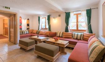 A cozy living room with a large L-shaped seating area and colorful cushions. Large windows let in plenty of light and the walls are cheerfully designed.