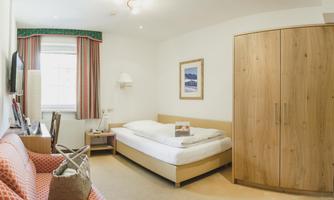 A simple room with a bed, sofa, and wooden wardrobe. There is a television and a window with curtains.