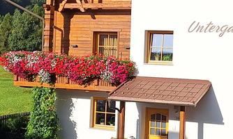 A cozy wooden house with a beautiful balcony full of flowers. In the background, green trees and a wide meadow are visible.