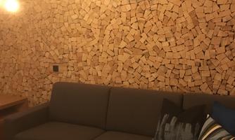 A cozy living room with a gray couch and a wooden table. The wall is decorated with wooden logs and warm lamps hang from the ceiling.