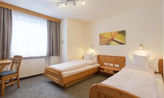A simple room with two single beds and a table. It has bright walls, carpeting, and large windows that let in plenty of light.