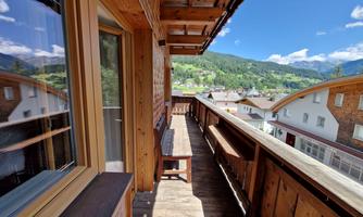 A spacious balcony with wooden cladding and a view of the mountains. The surroundings are green and inviting, with a blue sky and few clouds.