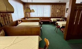 A cozy dining area with wooden furniture and tables covered with beige tablecloths. Large windows let in plenty of light and create an inviting atmosphere.