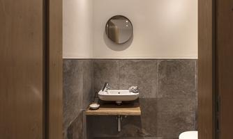 A modern bathroom with an elegant sink and a round mirror. The walls are made of gray tiles and there is a wooden shelf.