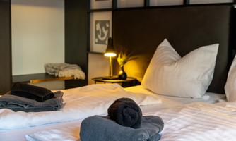 A cozy bedroom with a large bed and modern pillows. Neatly folded towels are placed on the bed, and a bedside lamp provides soft light.
