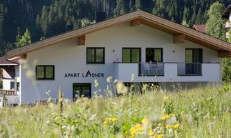 A modern building in the mountains with a green garden. The house has large windows and a balcony.