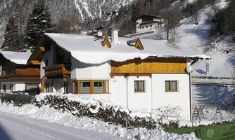 A charming house in the snow with wooden cladding and a beautiful balcony. In the background, snow-covered mountains and additional buildings are visible.