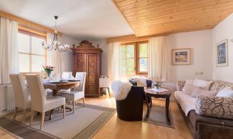 A cozy living space with wooden ceilings and large windows. There is a dining table, comfortable sofas, and decorative elements.