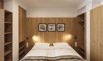 A modern bedroom with a double bed and warmer wooden furniture. Three pictures hang on the wall, and there are open shelves for personal items.