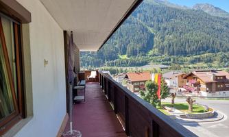 A balcony overlooking a green landscape and mountains. In the foreground, some houses and trees are visible.