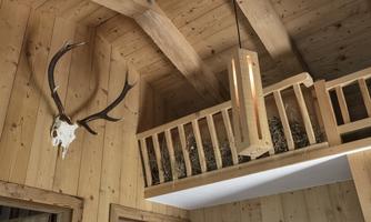 A cozy room made of wood with a modern wooden beam lamp. A deer antler hangs on the wall, and in the upper area, a small balcony can be seen.