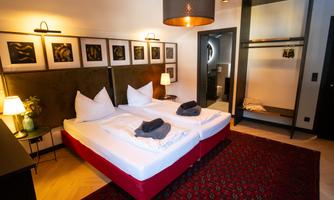 A modern bedroom with a double bed, decorated with white pillows and a red carpet. Artworks hang on the walls, and there is an open wardrobe.