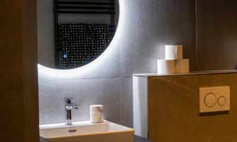 A modern bathroom with an elegant sink and stylish lighting. In the background, there is a decorative wall and toilet paper rolls visible.