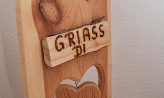 A wooden sign with the inscription "GRIASS DI" hangs on a wall. Below it is a heart-shaped cutout.