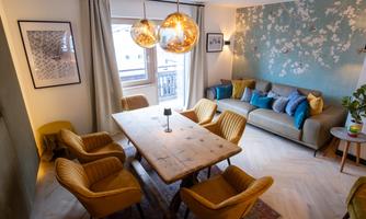 A modern living room with a large wooden table and yellow chairs. There is a floral wallpaper on the wall and a comfortable couch.